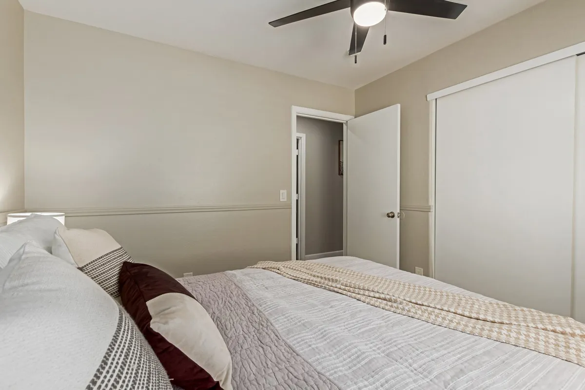 This bedroom image emphasizes a cozy bed situated near a window that lets in morning sunlight, complemented by a ceiling fan and a relaxing reading nook. The doorway hints at easy movement through the home.