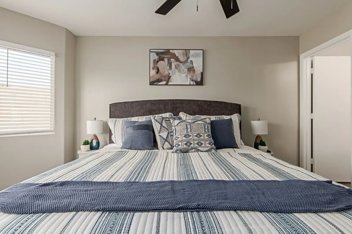 A spacious, inviting bedroom featuring a large bed with plush, patterned bedding and decorative pillows. The room is accented by stylish wall art and soft, warm lighting from table lamps, with natural light streaming in through windows. The overall mood is tranquil and cozy, perfect for restful nights or leisurely mornings.