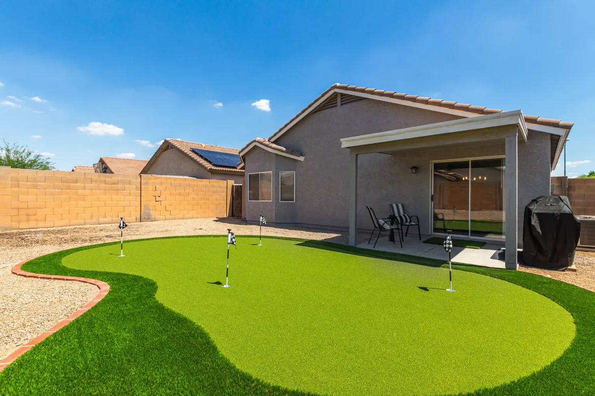 A panoramic angle of the backyard showing a manicured putting green, a welcoming patio area, and the rear view of the house. The image conveys ample space for gatherings and the secure, fenced nature of the yard.