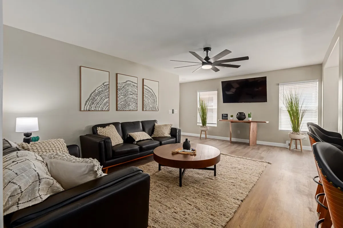The image showcases a bright, modern living room with plush black leather sofas arranged around a warm wooden coffee table, accented by vibrant indoor plants and striking wall art. A large TV mounted on the wall hints at cozy movie nights, while the inviting layout is perfect for relaxing, entertaining, or gathering with loved ones.