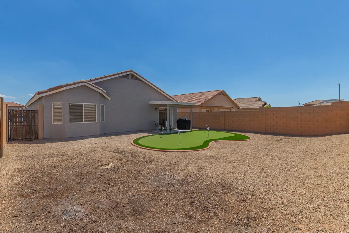 The image shows a putting green in the backyard, with the house facade and surrounding landscaping visible. The relaxed mood and open space suggest leisure and recreation, perfect for unwinding after a game.