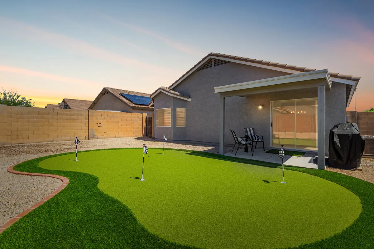 A backyard view showcasing the artificial putting green, patio seating area, and the home's exterior. The space is designed for outdoor relaxation, family gatherings, and playful activities, with sunset lighting enhancing the leisurely mood.