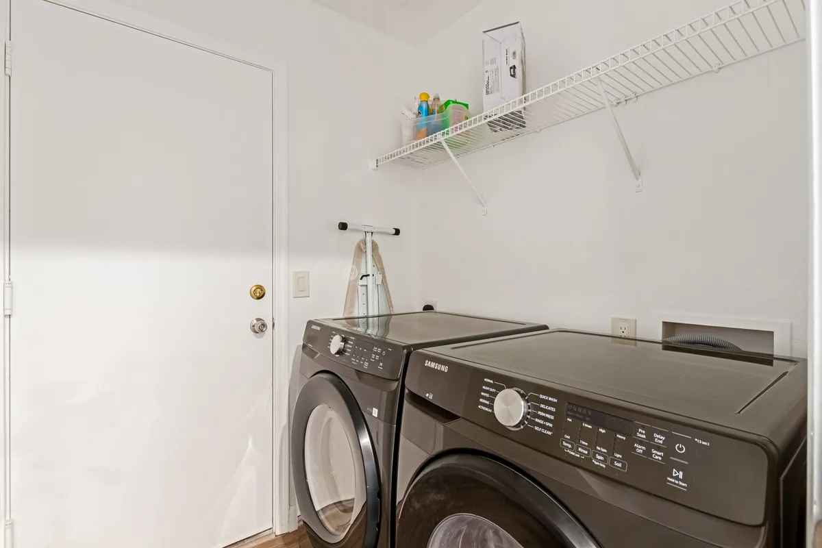 The image displays an in-unit laundry area featuring a front-loading washing machine and dryer, with shelves above containing cleaning supplies. The space feels orderly and accessible, making laundry straightforward for extended stays.